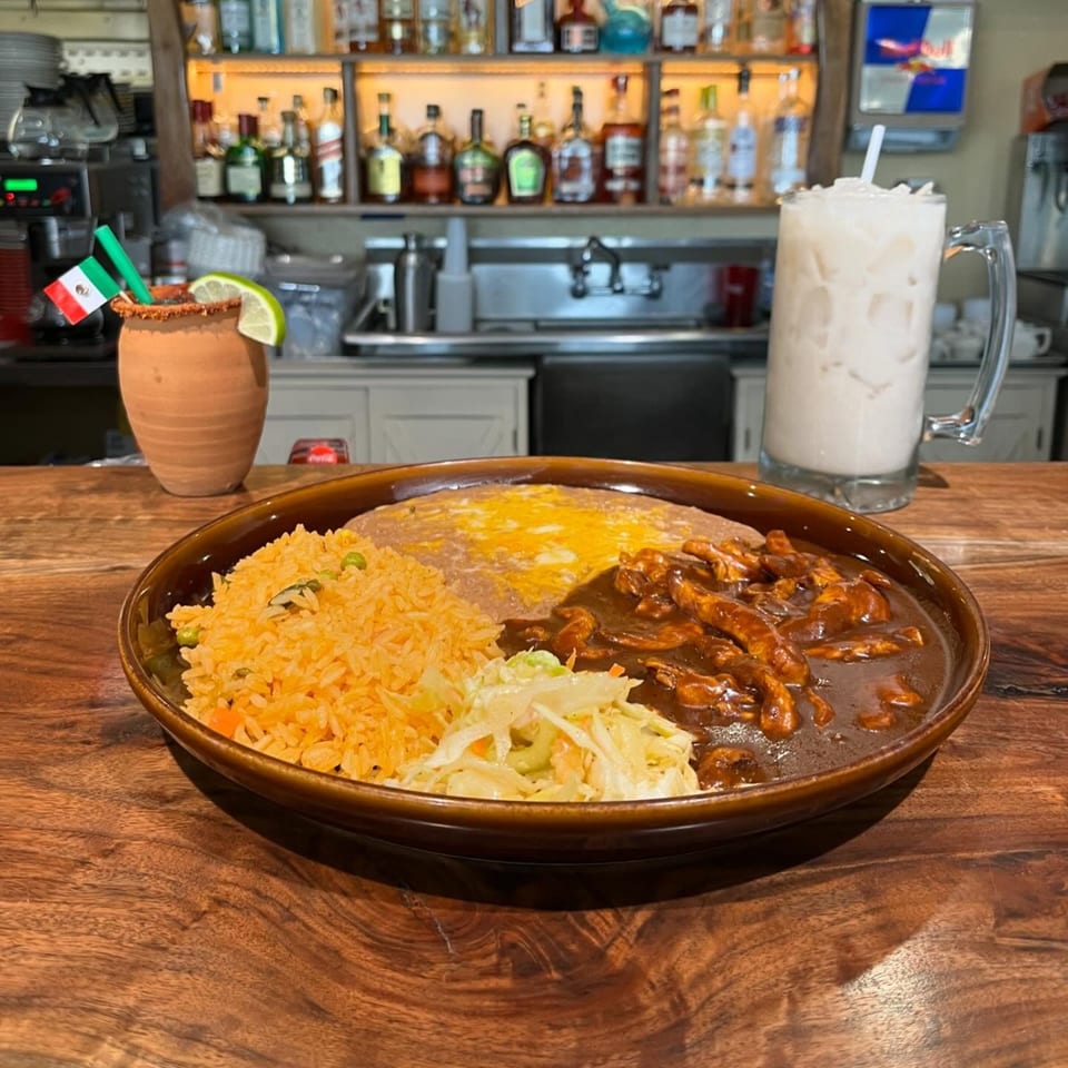 A Mexican dish with rice, beans, chicken mole, and coleslaw, with drinks in the background.