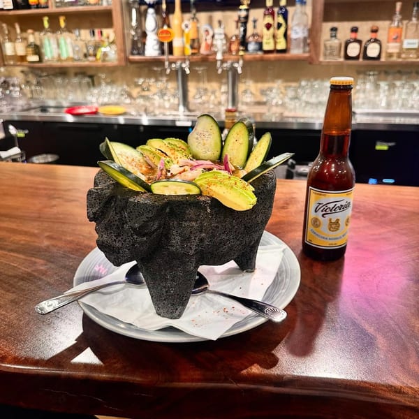 A molcajete filled with ceviche, avocado, and cucumber, next to a Victoria beer bottle at a bar.