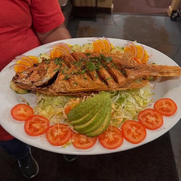 A whole fried fish, garnished with cilantro, served on a bed of lettuce with tomatoes, avocado, and citrus slices.