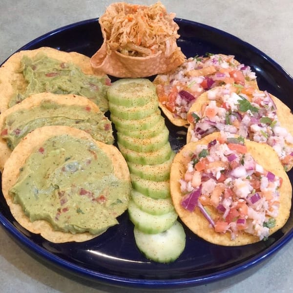 A blue plate with tostadas topped with guacamole and ceviche, alongside cucumber slices and a taco bowl.